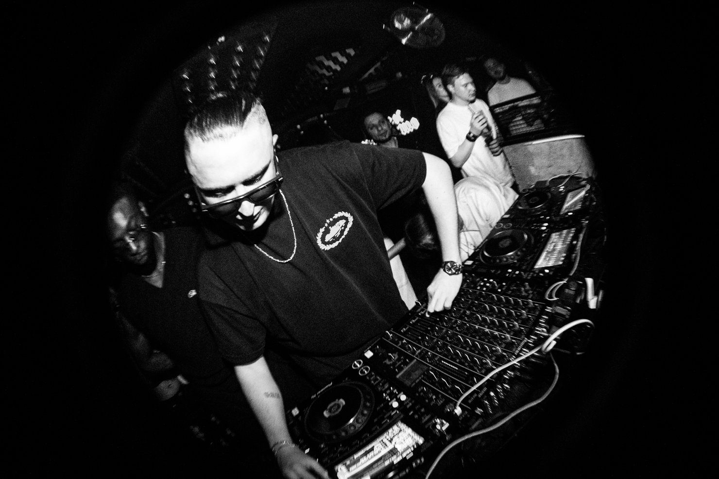 MNEEMO (Yaroslav Gorovoy) wearing glasses behind the DJ booth in a club, with a crowd in the background. Black-and-white photograph with an energetic atmosphere.