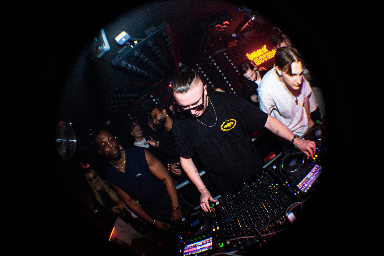 MNEEMO (Yaroslav Gorovoy) performing on the decks in a club, surrounded by people. The background is dark, and the lighting effects create a party atmosphere. A neon sign is visible.