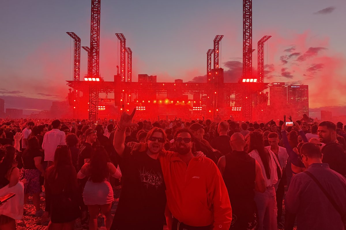Golden hour at Magazine Open-Air: the open-air stage rig with hanging line arrays, the Canary Wharf skyline behind the decks and thousands of people on the field, Greenwich Peninsula, 21 August 2026.