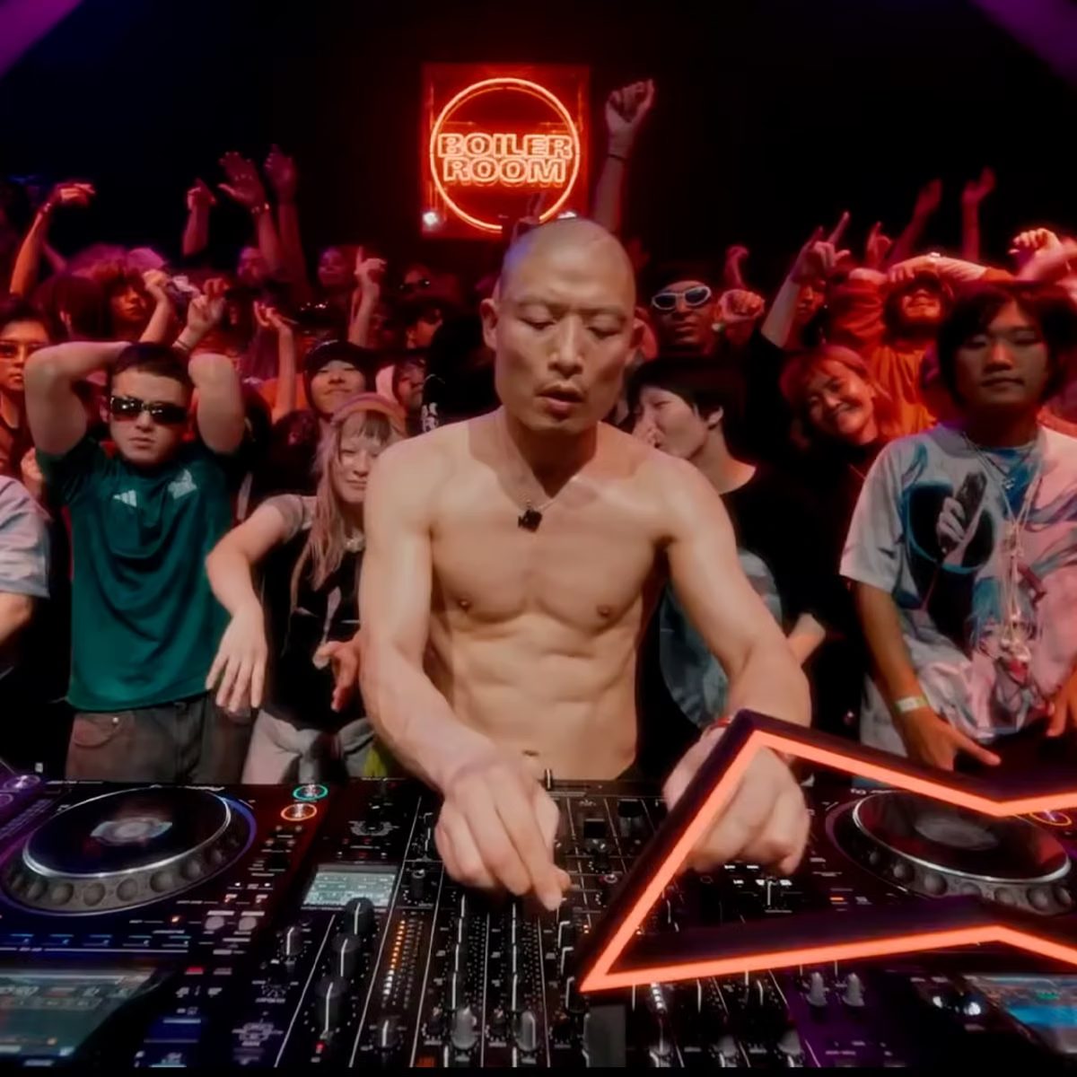 Yousuke Yukimatsu performing a Boiler Room set inside a packed dark club, surrounded by the crowd under red and purple lighting with the Boiler Room logo projected behind the booth.