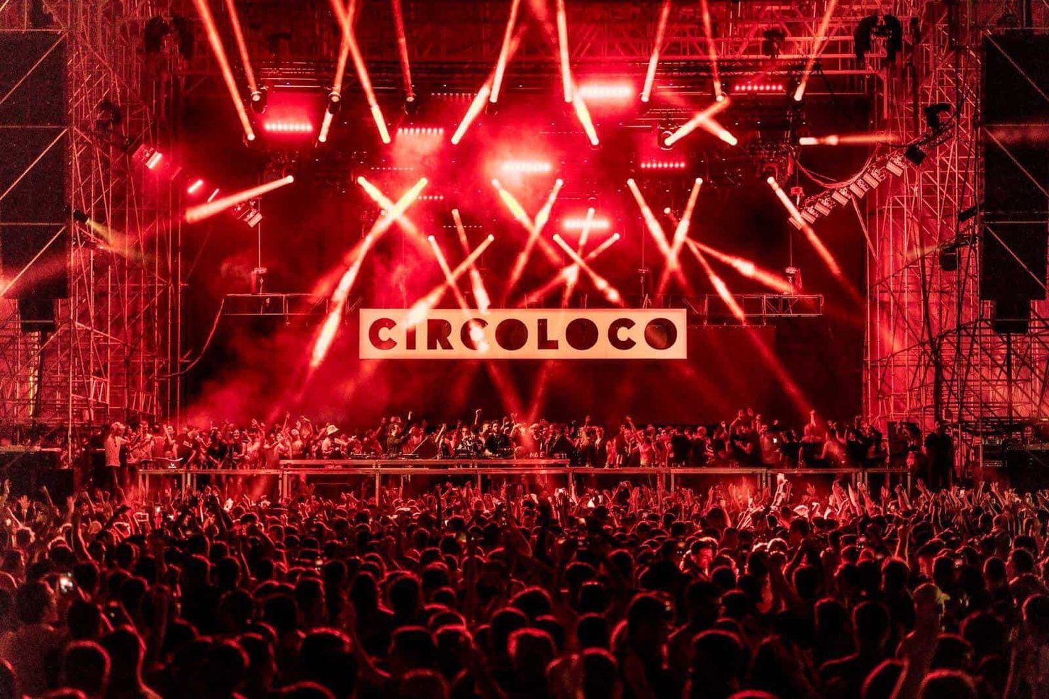 Crowd dancing under red lights at a Circoloco event. The stage is filled with people, and beams of light illuminate the scene, creating a vibrant atmosphere.