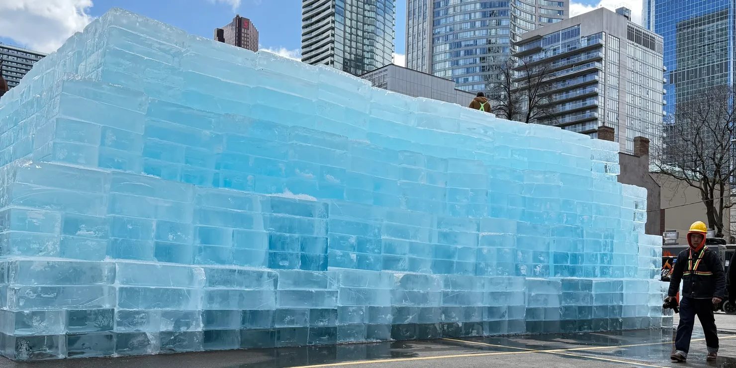 Drake Iceman concept art. Blue ice wall in a city setting, with skyscrapers in the background. A worker in a yellow hard hat walks nearby. Clear, sunny day.