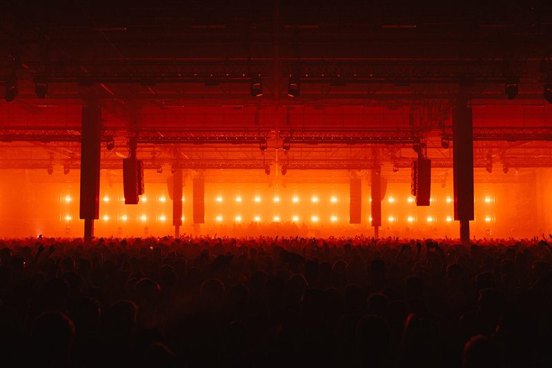 Drumsheds 2026. A large crowd at a concert in a dimly lit hall with bright orange lights. The atmosphere is energetic and dynamic.