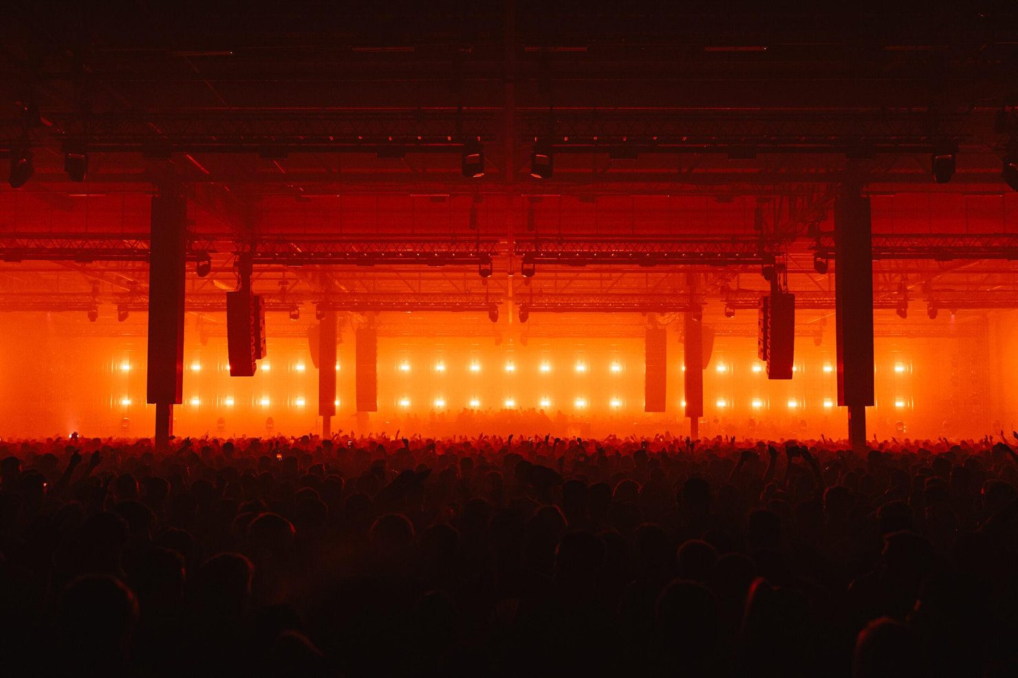 Drumsheds 2026. A large crowd at a concert in a dimly lit hall with bright orange lights. The atmosphere is energetic and dynamic.
