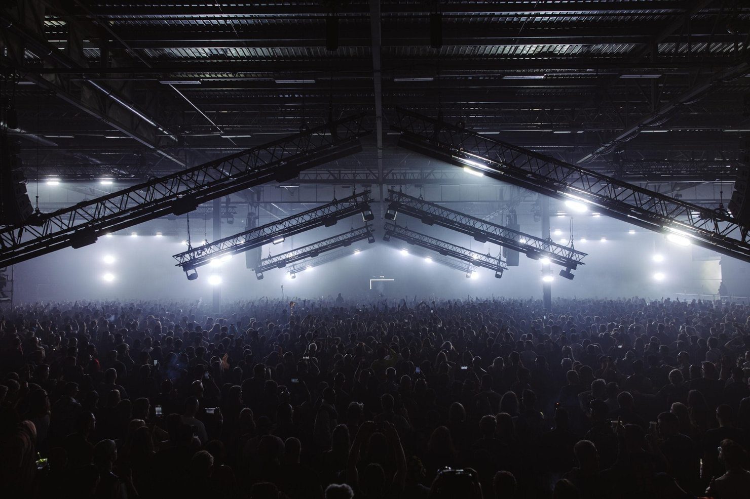 Drumsheds 2026. Crowd fills a large, industrial-style venue with overhead lighting and fog. Mood is lively and energetic with beams and spotlights.