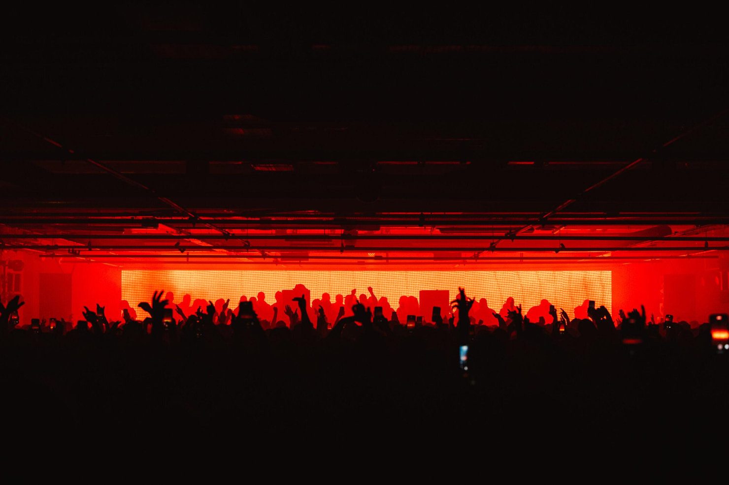 Drumsheds 2026. Silhouettes of a crowd at a concert under vivid red lighting. People raise hands and phones, creating an energetic, lively atmosphere.