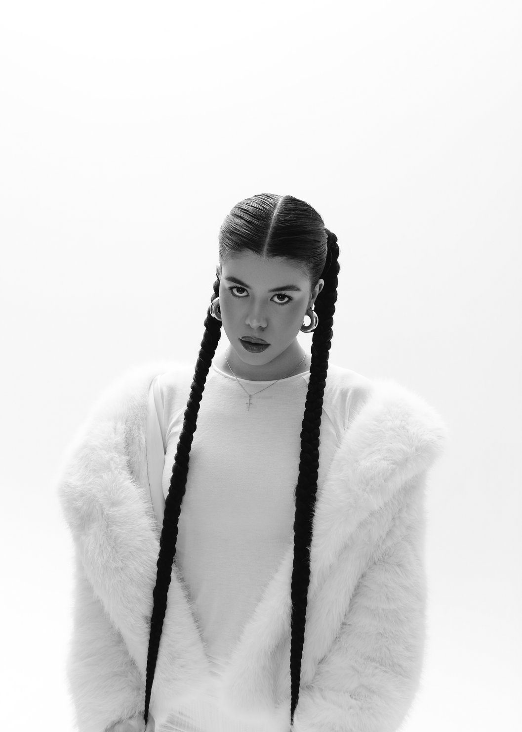 emi (Emilie Arfush) with long braids and earrings, wearing white fluffy clothing, against a grey background. A serious mood.
