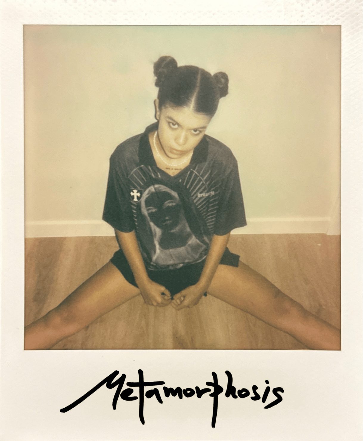 emi (Emilie Arfush) with hair in buns, sitting on the floor with her legs apart. An icon is printed on the T-shirt. Light-coloured floor, with the text “Metamorphosis” at the bottom.