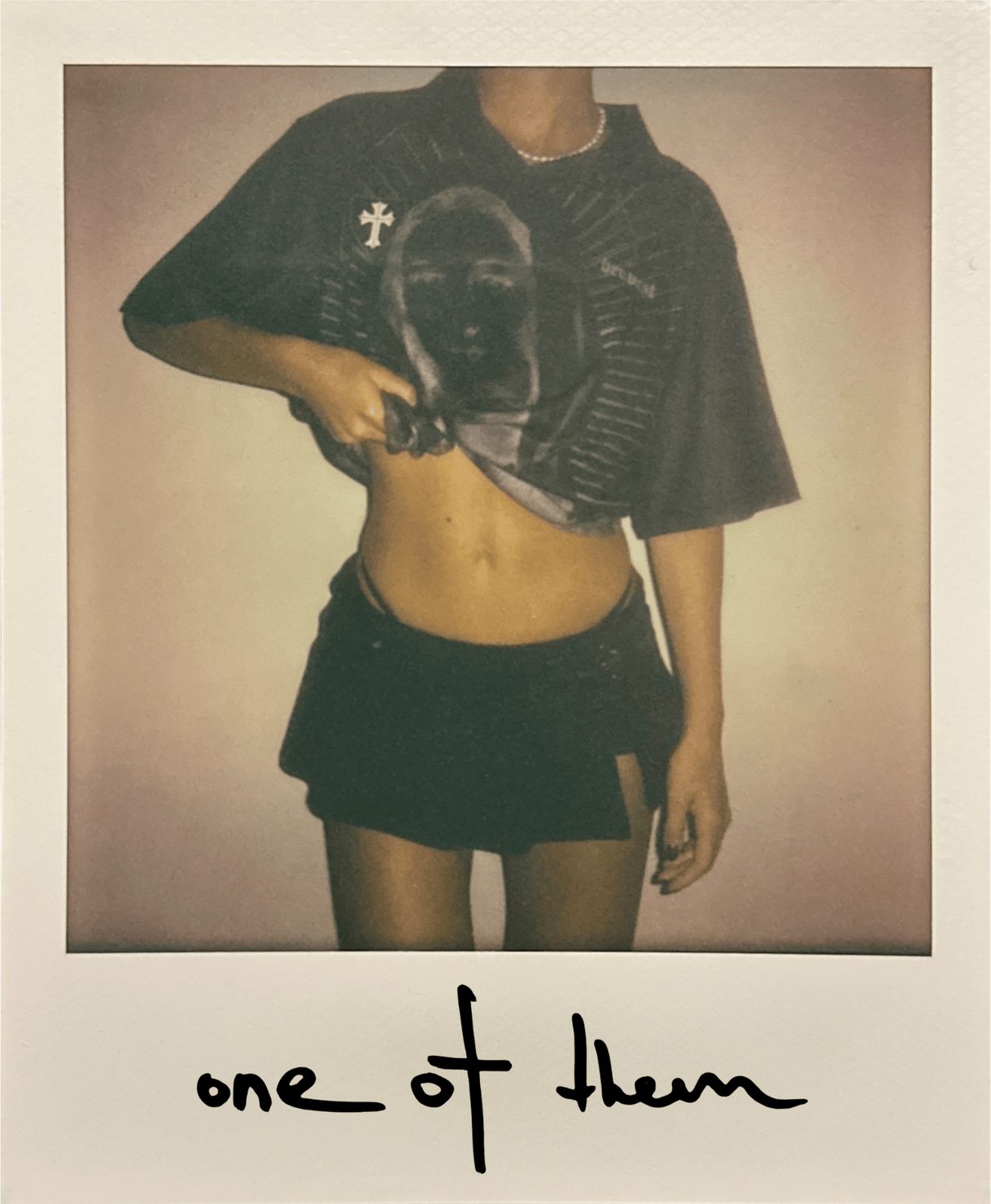 emi (Emilie Arfush) wearing a T-shirt with a graphic print, lifting it slightly to reveal her stomach. Caption: “one of them”. Light-coloured wall in the background.