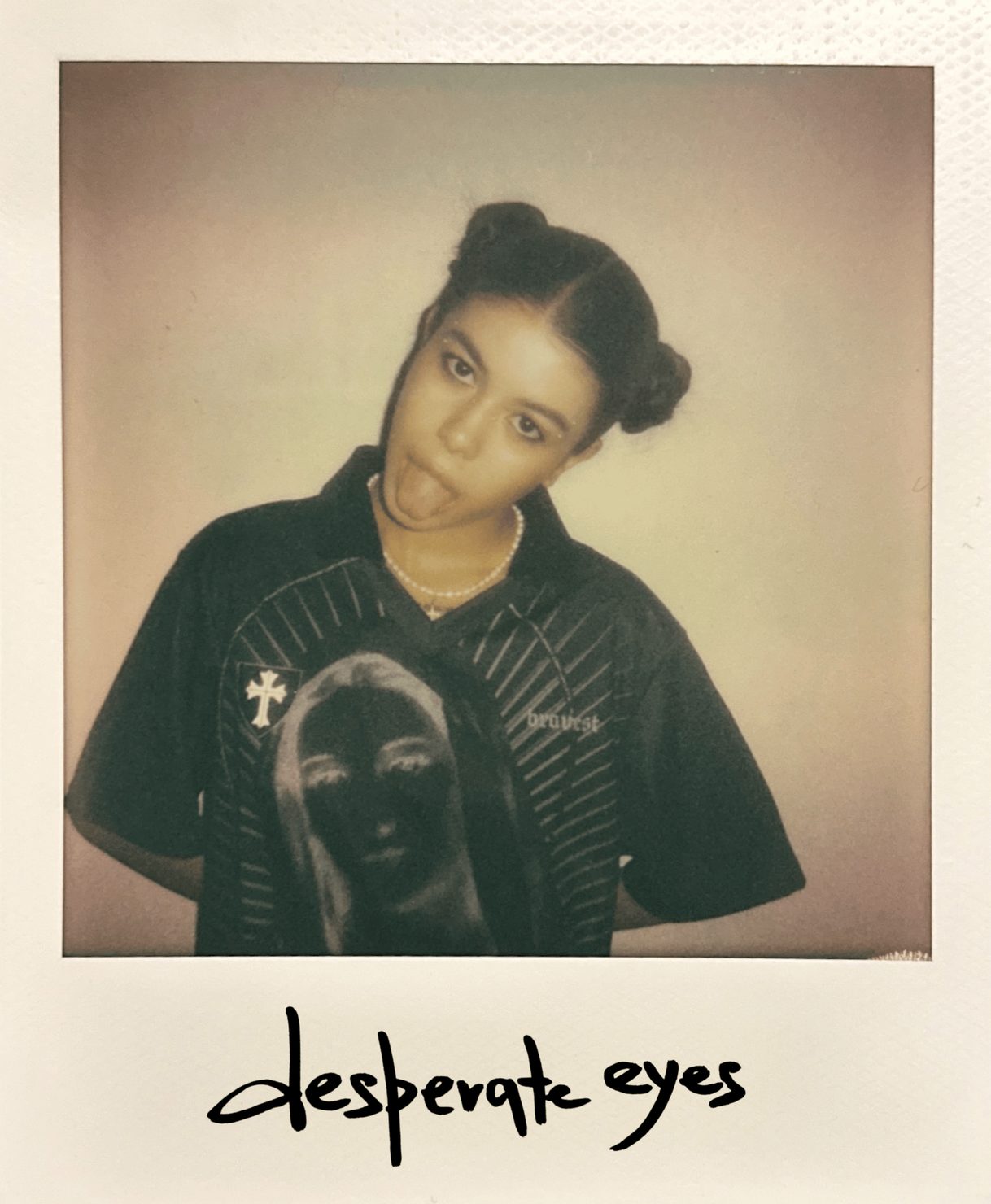emi (Emilie Arfush) with two buns, sticking her tongue out. An image is printed on the T-shirt, with a necklace around her neck. Text: “desperate eyes”. Neutral background.