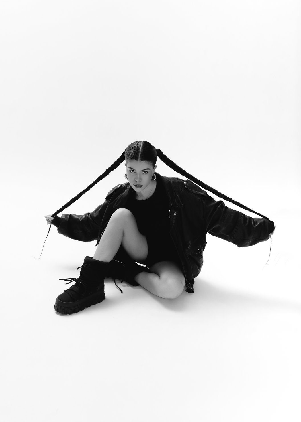 emi (Emilie Arfush) in black clothing, sitting on the floor in a studio, holding her long braids out to the sides. Black boots, earrings. Black-and-white photo.