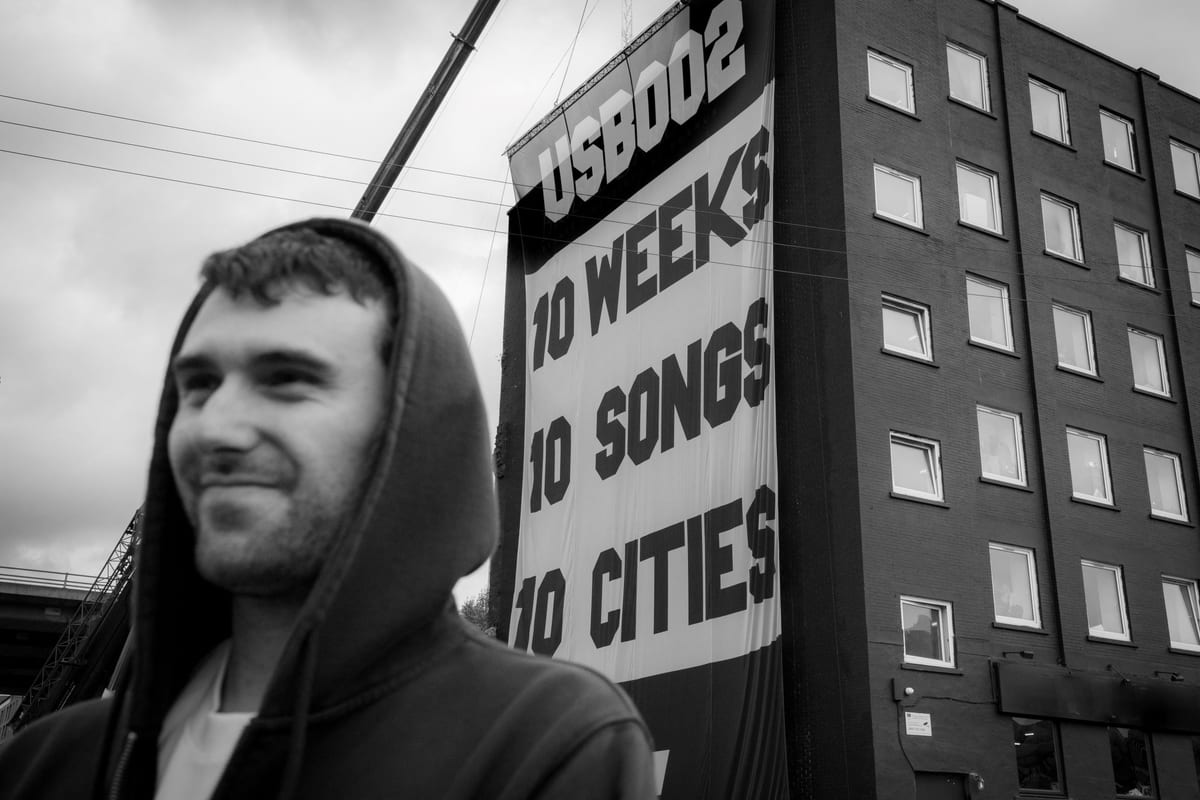 Fred again.. in front of the USB002 campaign banner reading 10 weeks, 10 songs, 10 cities, the secret-show concept that defined his 2025 to 2026 run
