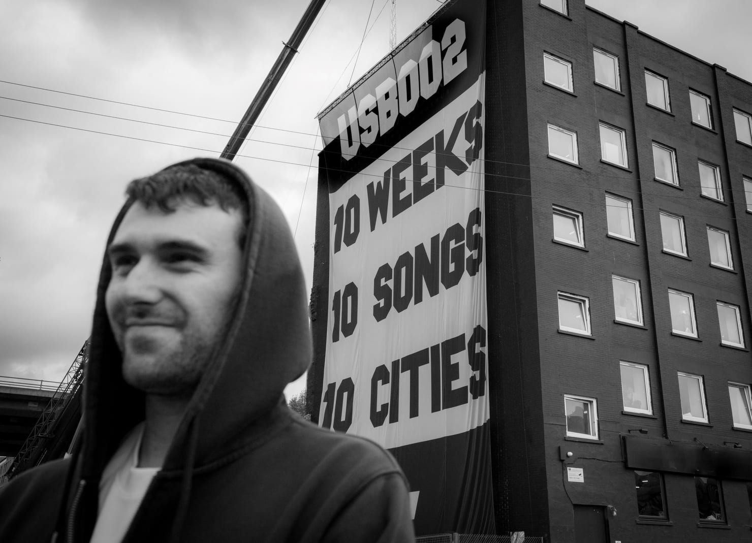 Fred again.. in front of the USB002 campaign banner reading 10 weeks, 10 songs, 10 cities, the secret-show concept that defined his 2025 to 2026 run