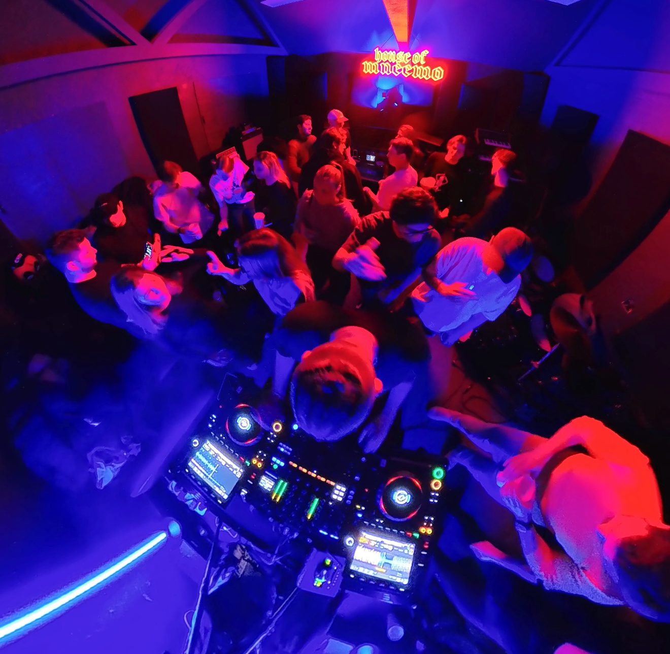 MNEEMO (Yaroslav Gorovoy) DJing for dancing people under neon blue and red lighting. On the wall behind them, a glowing sign reads “house of mneemo”.
