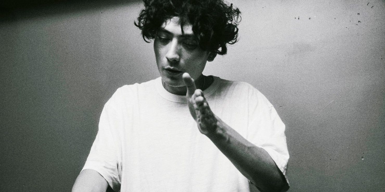Interplanetary Criminal in a white T-shirt, with curly hair, making a hand gesture and looking down. Black-and-white background, calm expression.