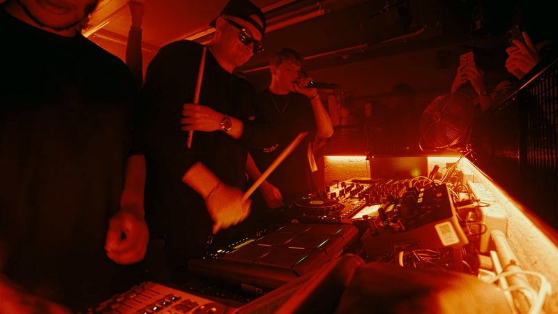 MNEEMO (Yaroslav Gorovoy) with drumsticks and singer MJP (Matthew John Potter) perform in a dimly lit club, surrounded by equipment and recording onlookers. Red lighting sets the energetic mood.