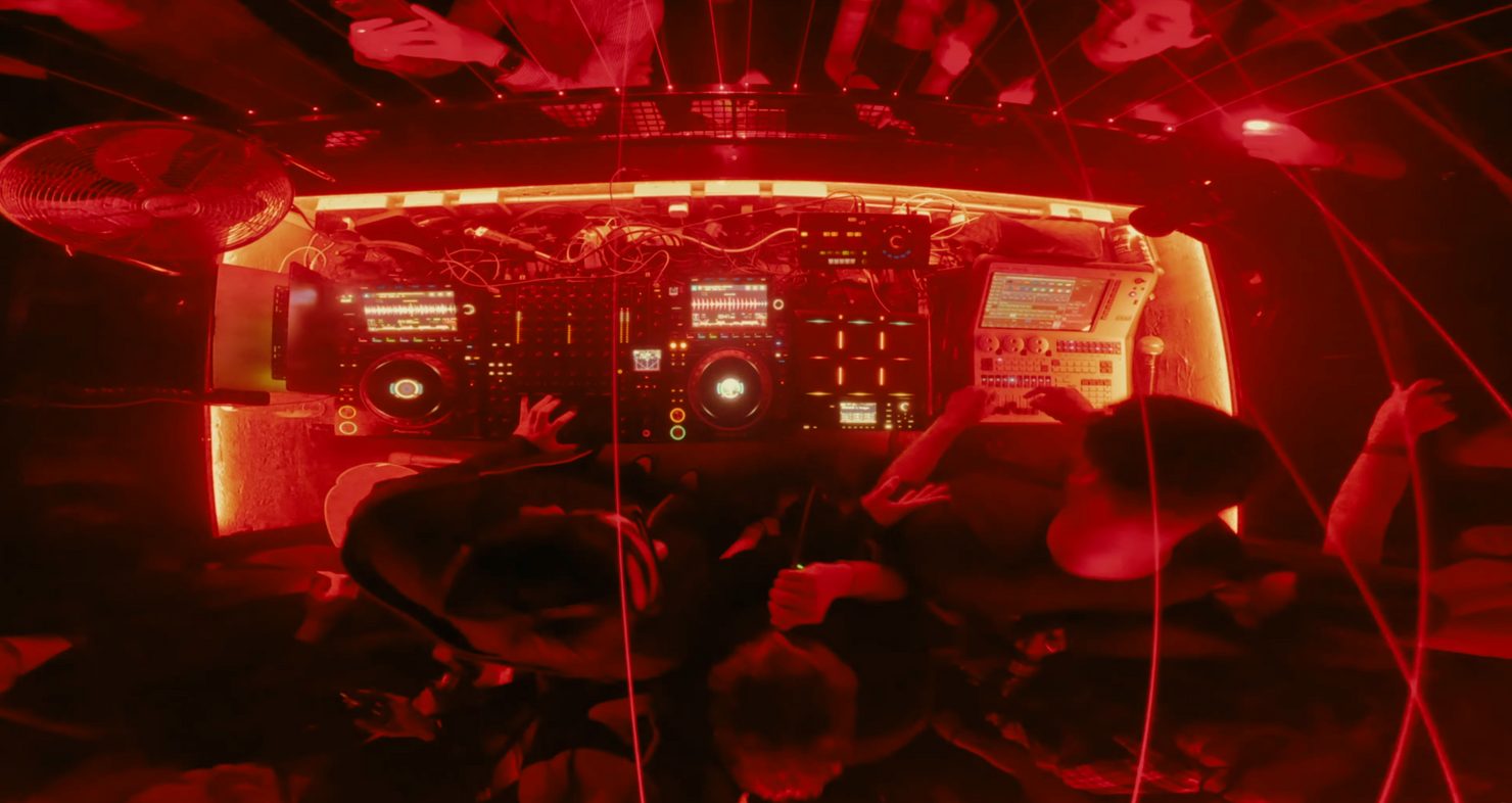 DJ booth in a club with people dancing. Red lighting, digital mixers, and screens create an energetic atmosphere. MNEEMO (Yaroslav Gorovoy) DJing in the middle.