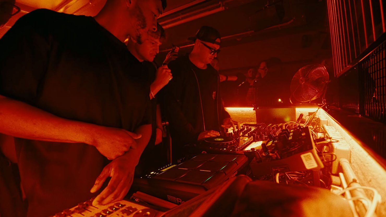 MNEEMO (Yaroslav Gorovoy) performing with equipment in dimly lit room, red lighting creates an intense atmosphere. Crowd in background watches closely.