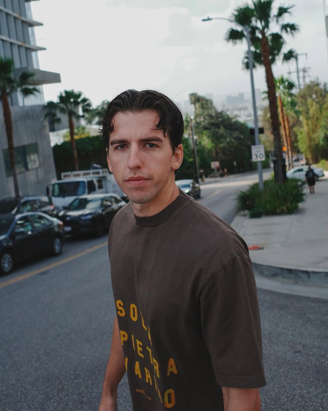 Josh Baker standing on a palm-lined Los Angeles street, looking directly at the camera in a relaxed streetwear outfit.