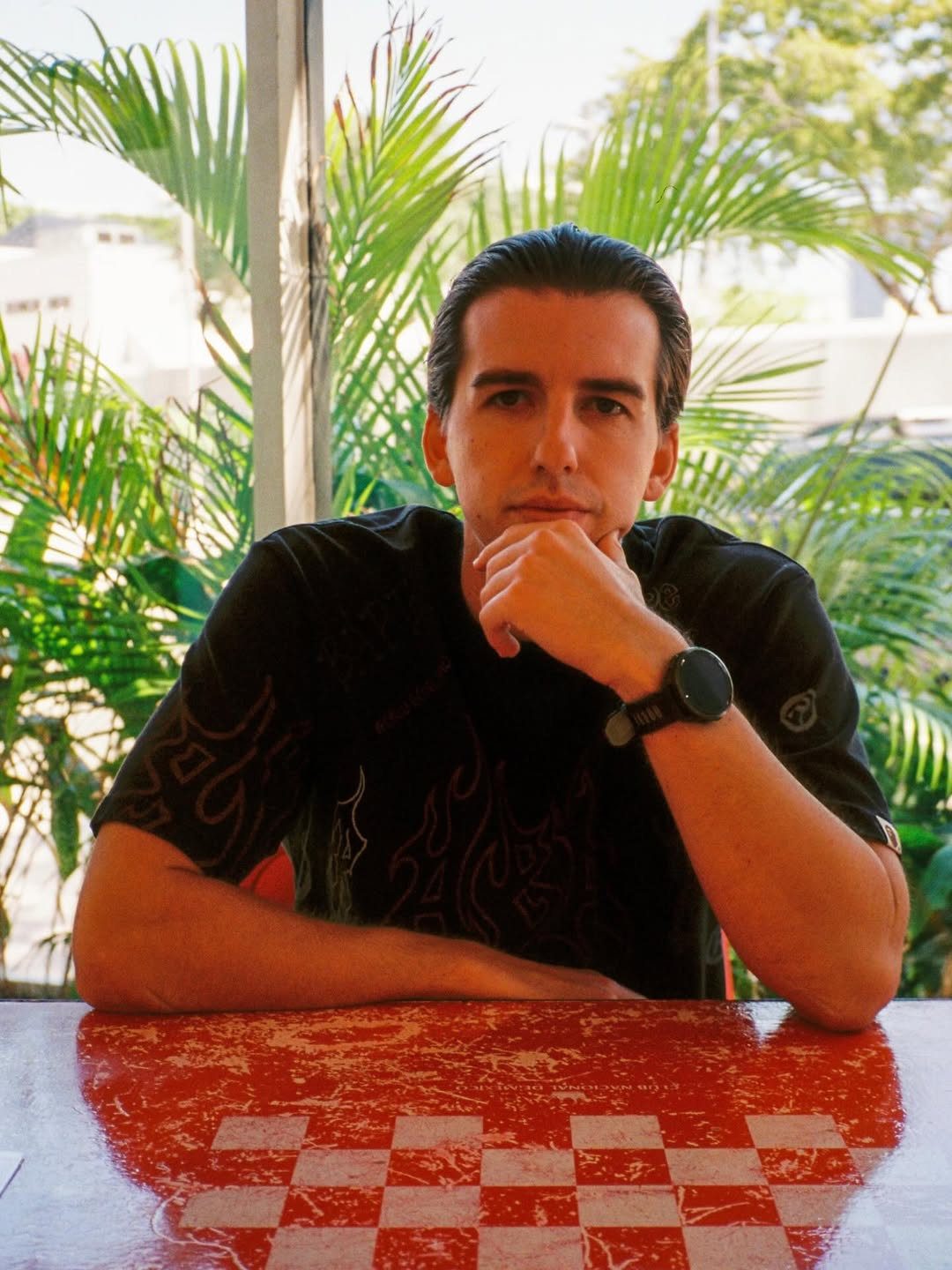 Josh Baker seated at an outdoor table surrounded by tropical plants, looking into the camera with a calm, focused expression.