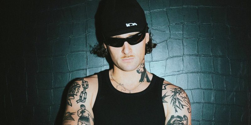 KETTAMA wearing a black beanie and sunglasses, standing against a dark wall. Tattoos visible on the body, with a serious expression.