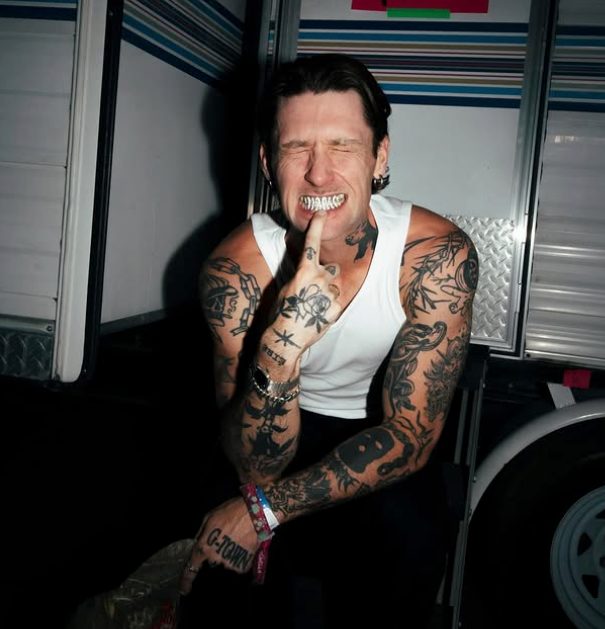 KETTAMA Real Name Evan Campbell in a white tank top shows off grills, smiling with eyes closed. He's seated, with colorful wristbands and trailer in the background.