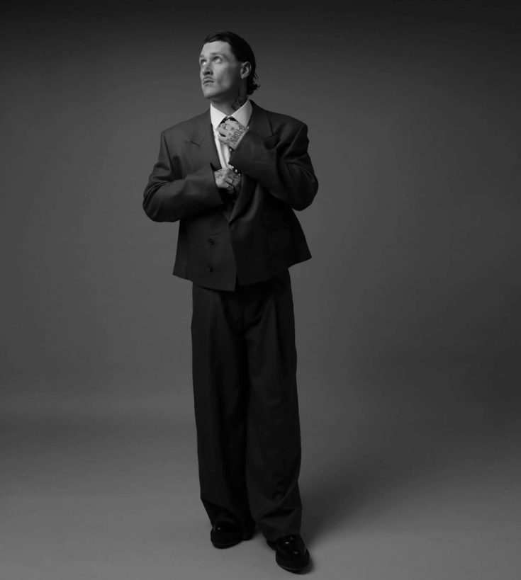 KETTAMA Real Name Evan Campbell in oversized suit adjusting tie, looking thoughtful. Black-and-white image with a neutral studio background.