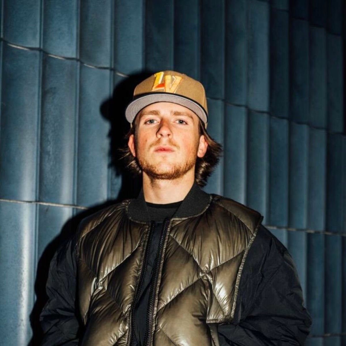 Luke Dean wearing a jacket and an “LV” cap, posing against a blue tiled wall. Calm expression.