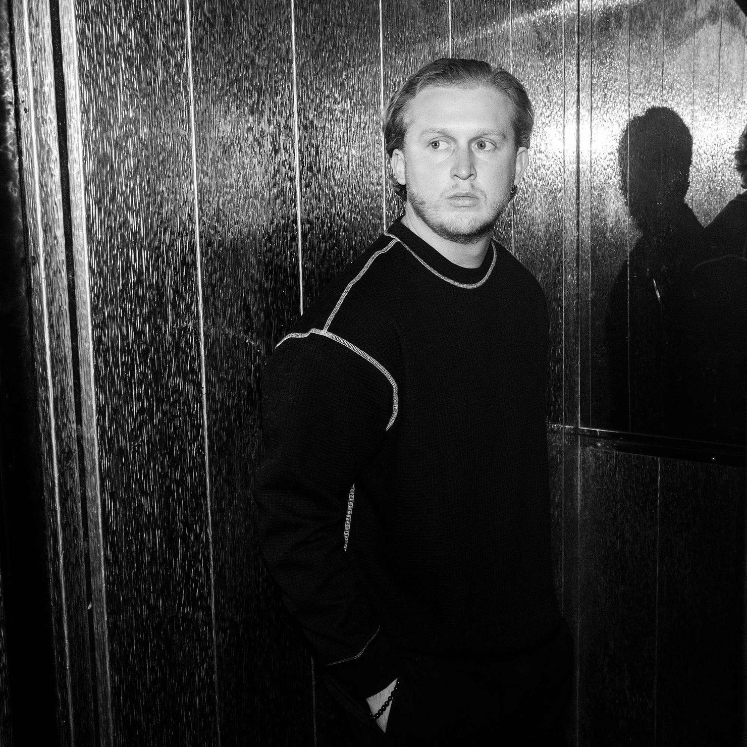 Max Dean in dark clothing, standing by a wall and looking into the distance. Black-and-white palette, reflection in the glass, thoughtful atmosphere.