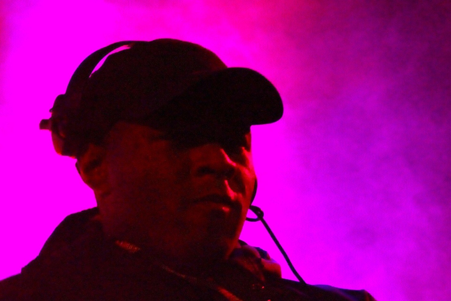 DJ EZ in a black cap and headphones, lit magenta on stage, looking out over the crowd