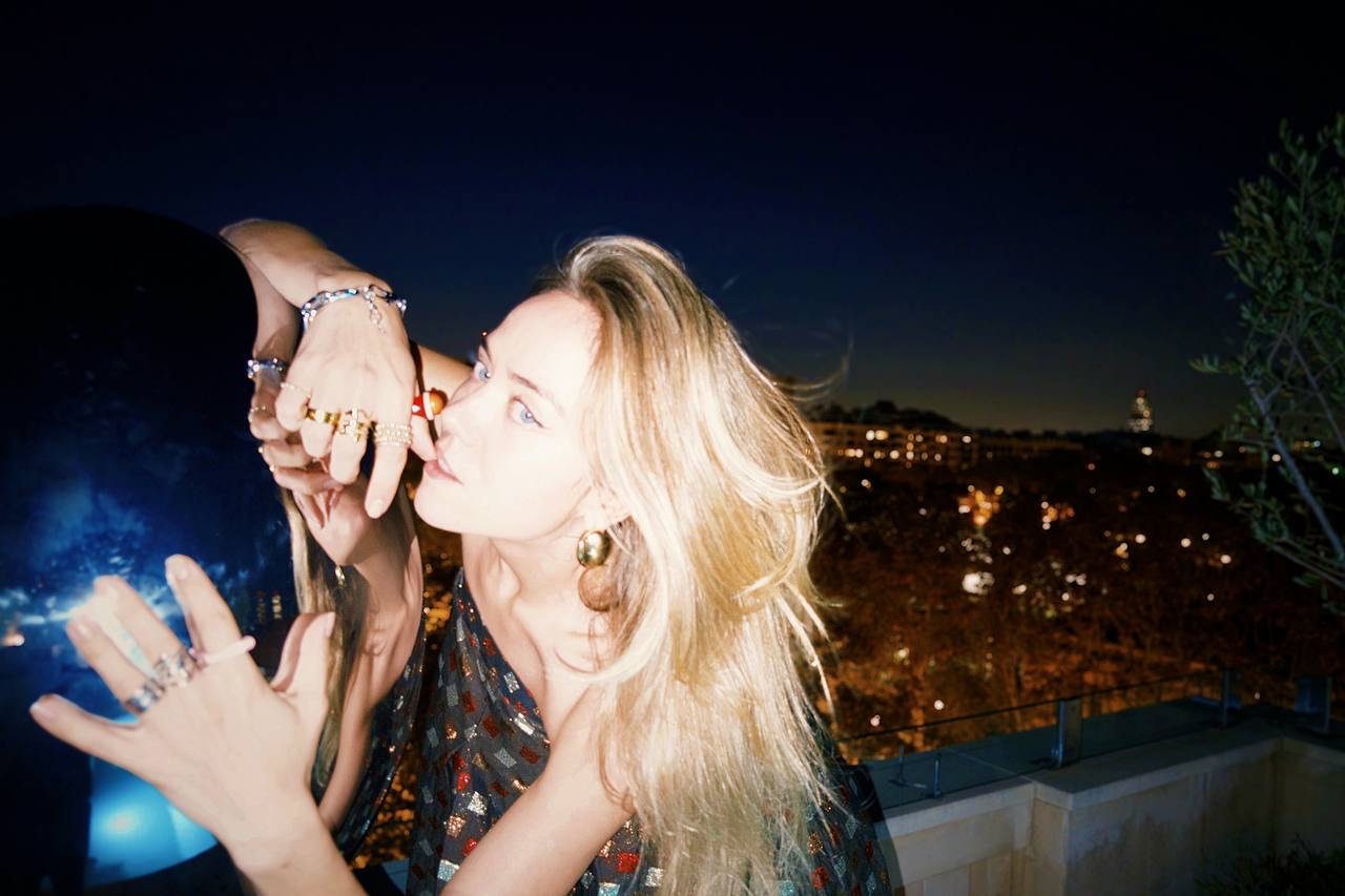 Elizabeth Lip with long blonde hair against a night-time city backdrop. She is wearing a sparkling dress and jewellery, looking into the camera with a thoughtful expression.