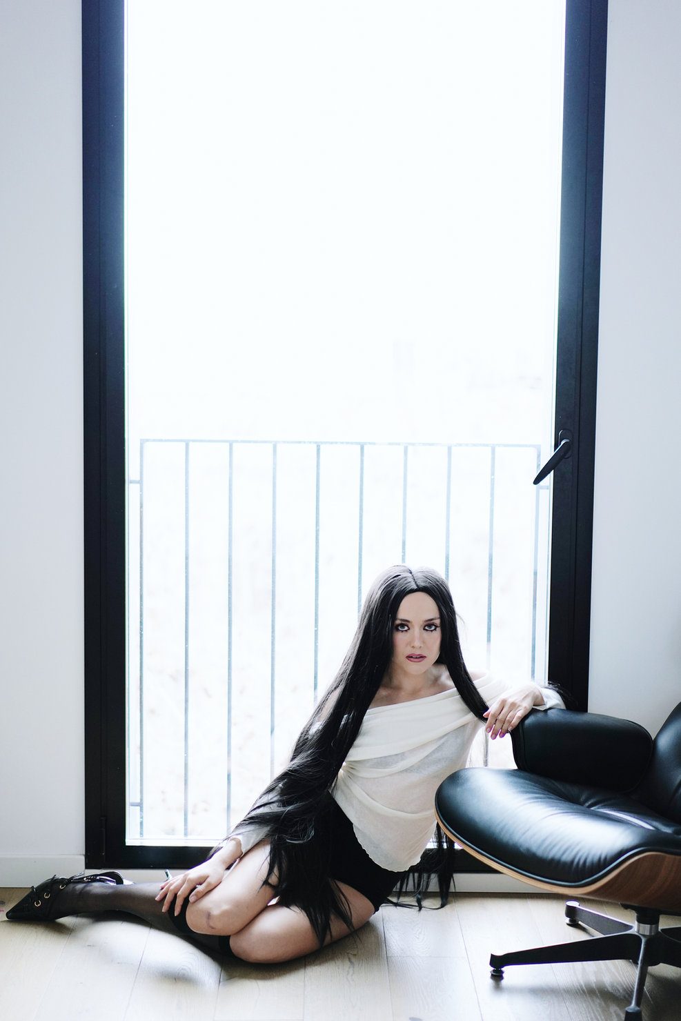 Человек Elizabeth Lip during the promo campaign for “Slow”, wearing a white blouse and a black skirt, sitting on the floor by a window. Long black hair, thoughtful gaze. A black leather armchair beside her.