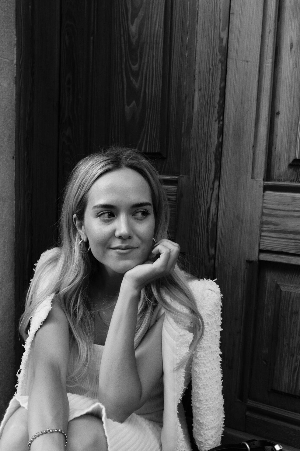 Elizabeth Lip with long blonde hair, wearing earrings and a bracelet, smiling thoughtfully while sitting by a wooden door. Black-and-white photo.