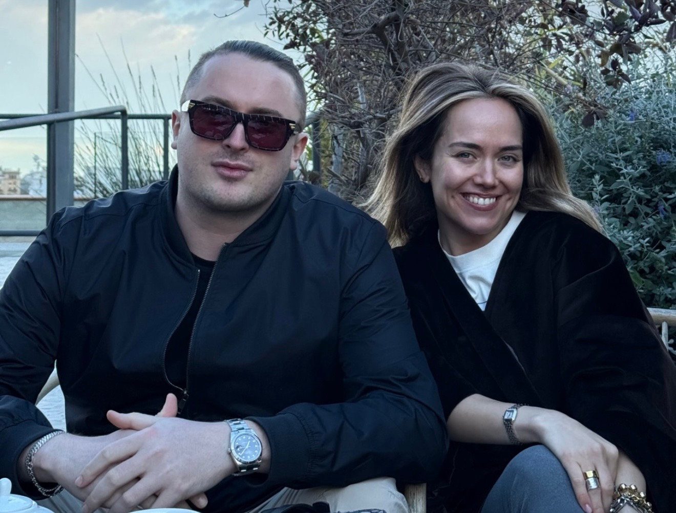 Elizabeth Lip & MNEEMO (Yaroslav Gorovoy) sitting outdoors, smiling. The man is wearing a dark jacket and glasses, and the woman is in a black coat. Background: greenery and railings.