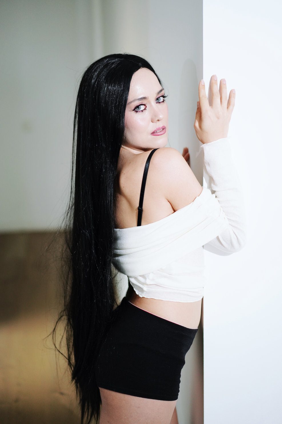 Elizabeth Lip during the promo campaign for “Slow”, with long black hair, wearing a white top and black shorts, standing by a wall and looking back over her shoulder. Light background, calm expression.