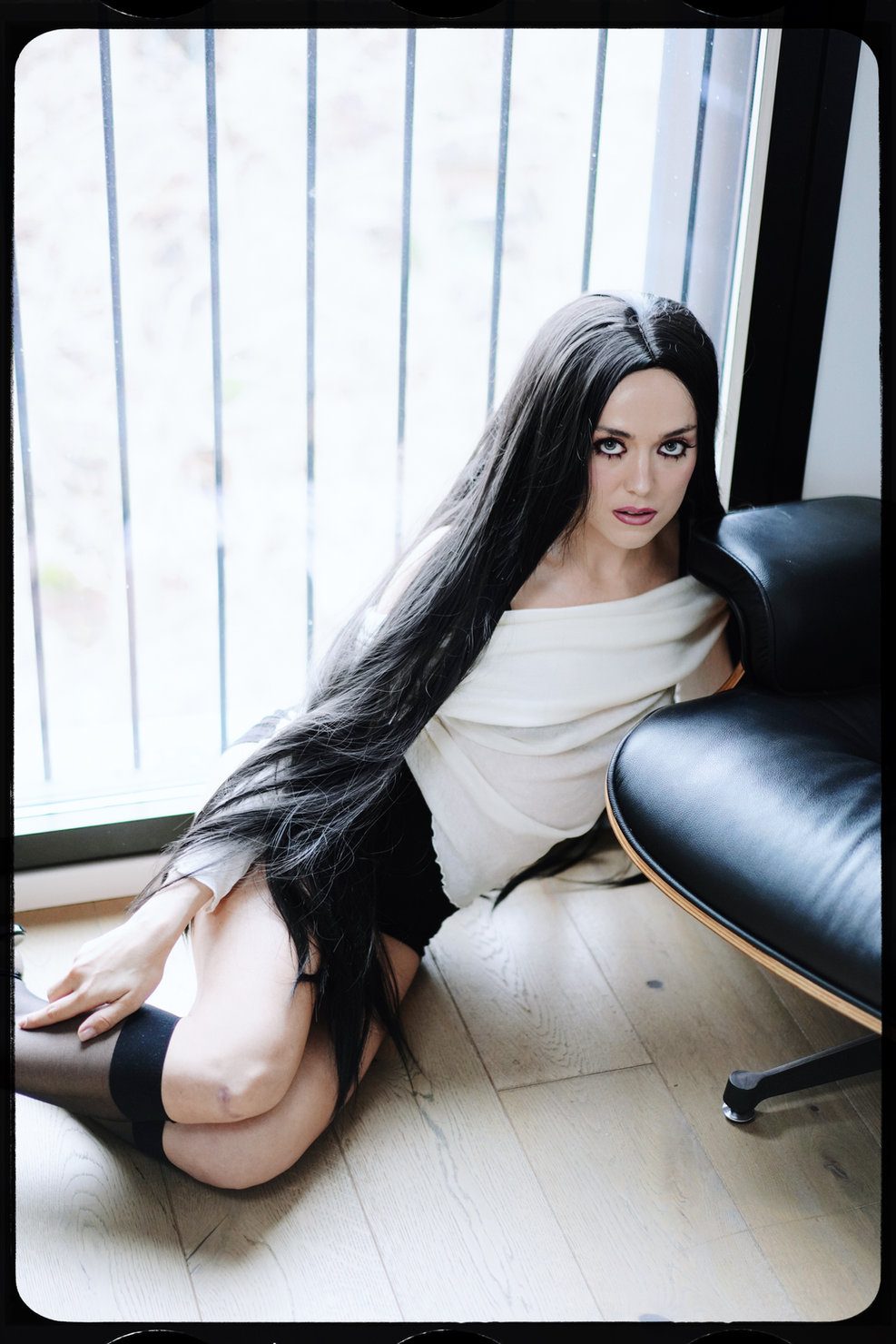 Elizabeth Lip during the promo campaign for “Slow”, with long black hair and a serious expression, sitting on the floor by a window. Light-coloured dress, dark leather furniture.