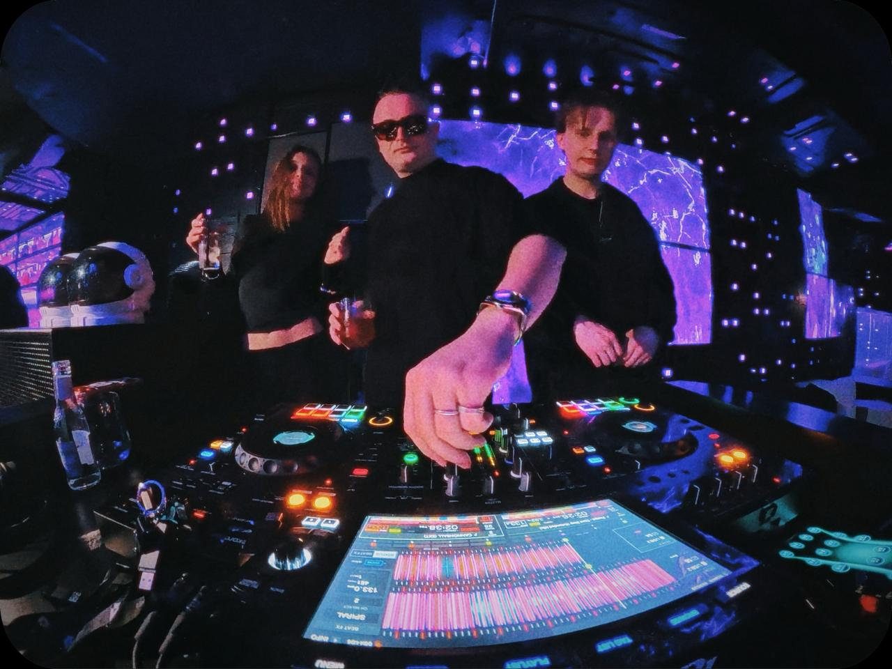 MNEEMO (Yaroslav Gorovoy) in a club behind the DJ booth, with FLASH & Ryeli beside him holding drinks. Purple lights create the atmosphere. Bright buttons and a screen are visible.