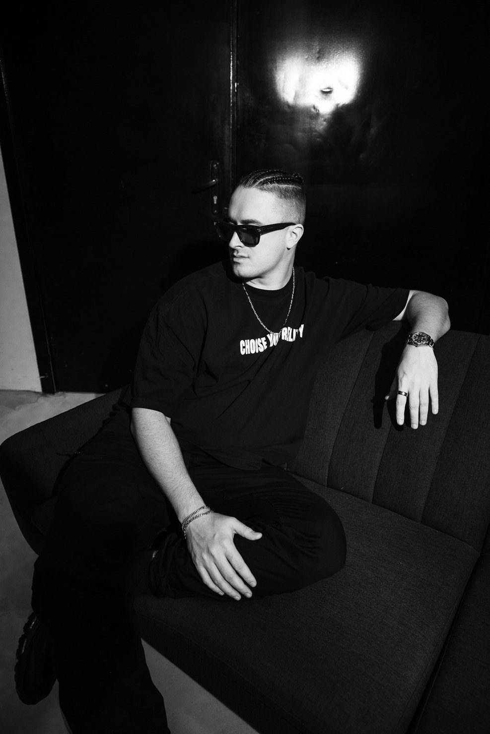 Мужчина MNEEMO (Yaroslav Gorovoy) wearing black sunglasses, sitting on a sofa. He is dressed in a black T-shirt with the text “CHOOSE YOUR REALITY” by APTEKA24, posing in a dimly lit room.