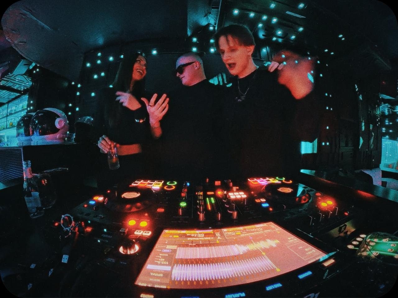 MNEEMO (Yaroslav Gorovoy), FLASH & Ryeli by the DJ booth: one is singing, the woman is laughing, and the other is wearing glasses. Blue lighting in the background, with a fun, music-filled atmosphere.