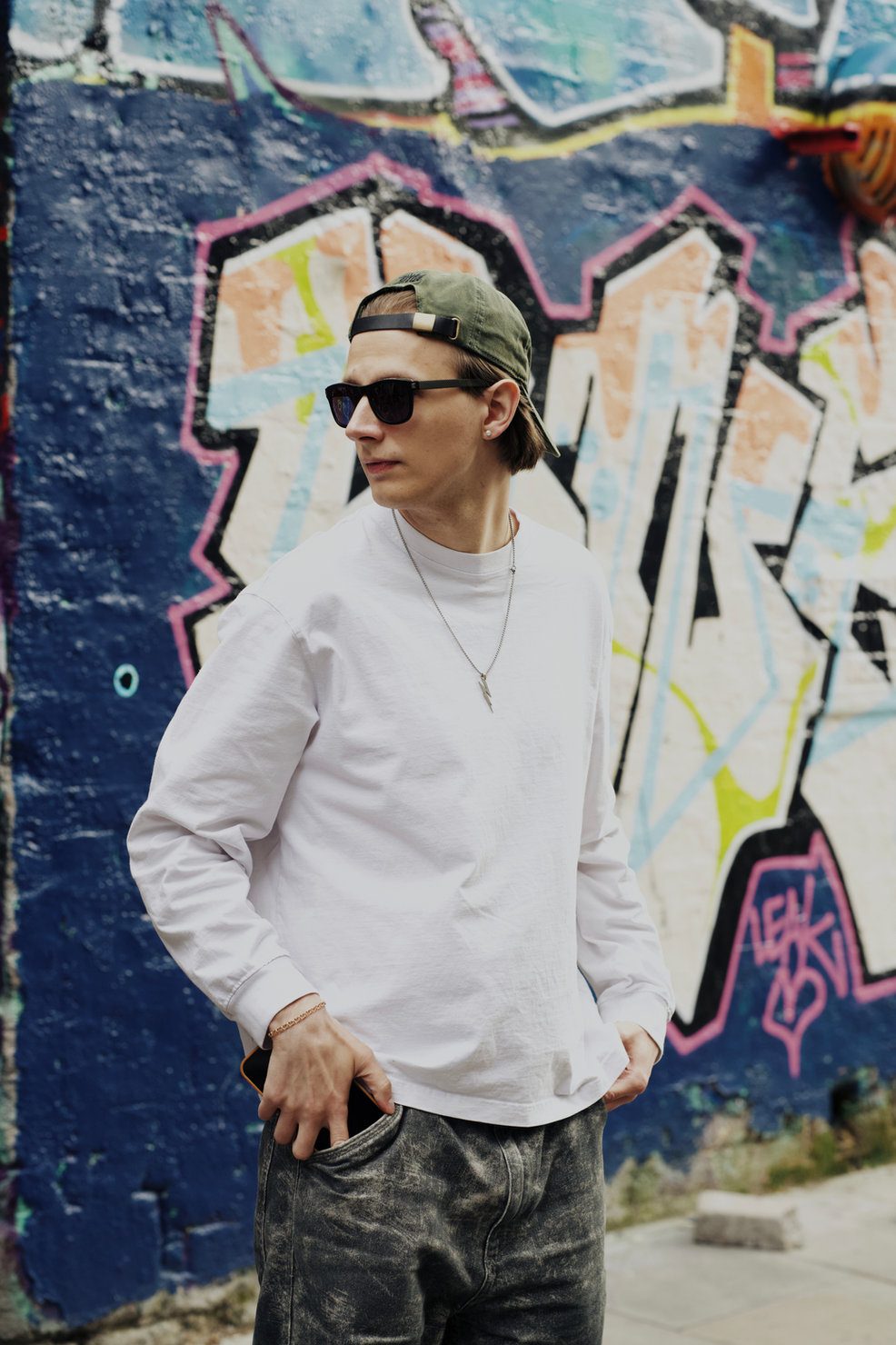 FLASH (Anton Ditiashov) wearing a white sweatshirt and sunglasses, standing against a graffiti-covered wall. With one hand in his pocket, he looks relaxed.