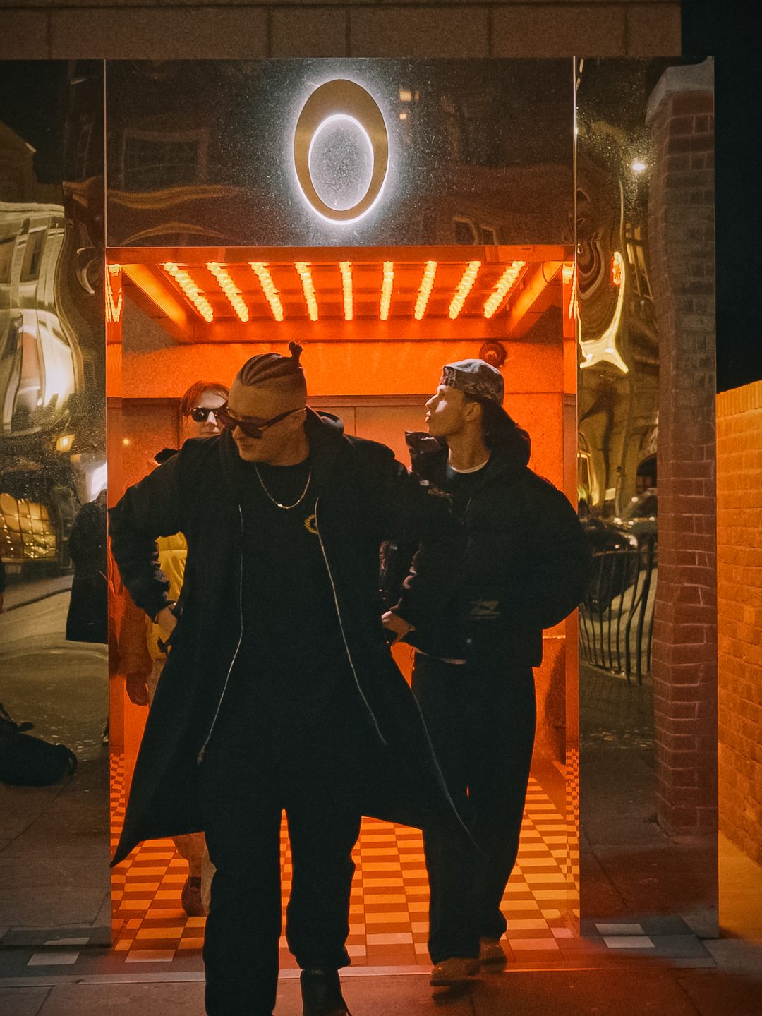 MNEEMO (Yaroslav Gorovoy) and friends in black clothing, walking out of a building lit with orange light. A round illuminated sign hangs above the entrance. Night-time city atmosphere.