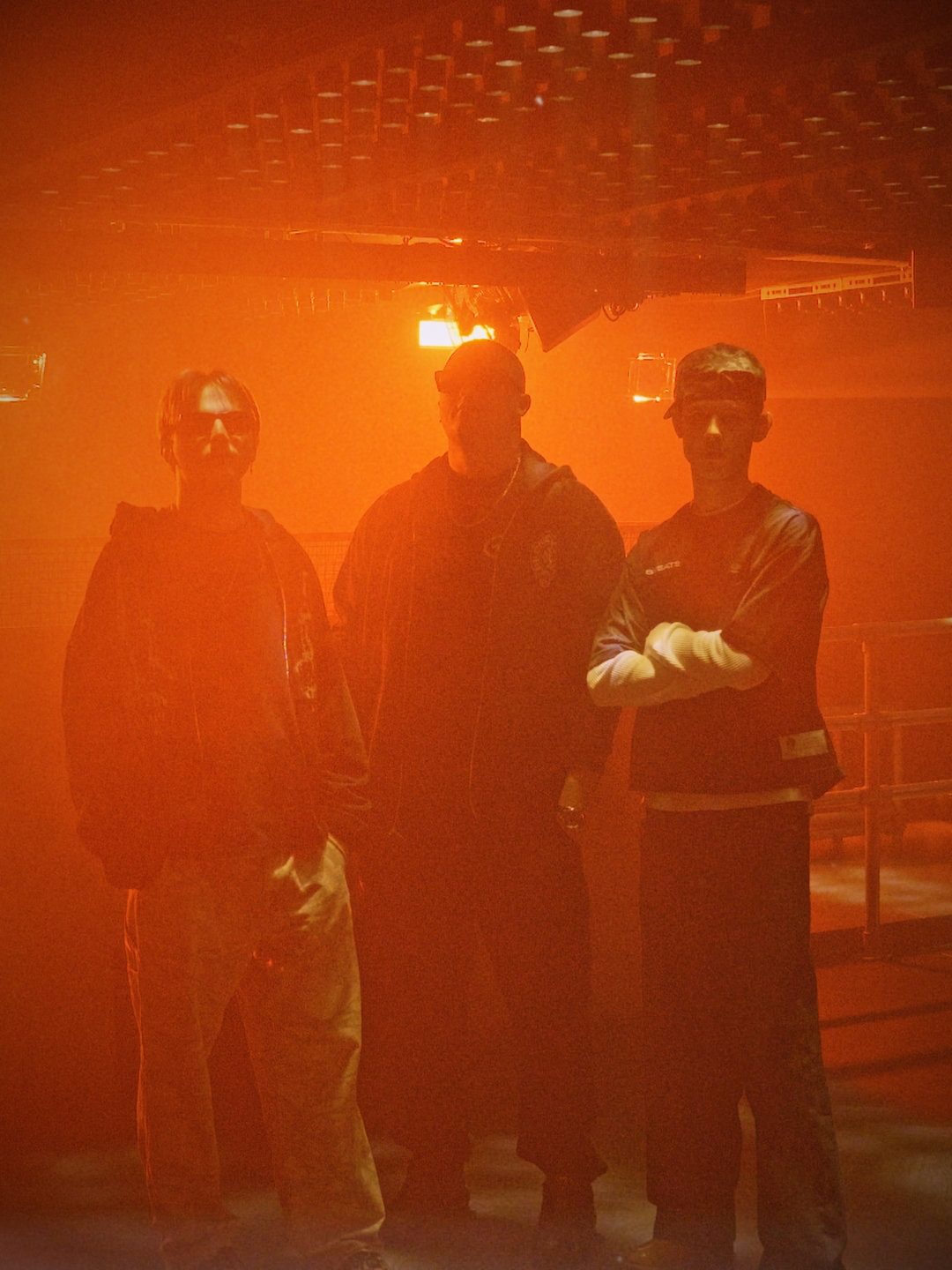 MNEEMO (Yaroslav Gorovoy) and friends standing in a club with orange lighting. All dressed in dark clothing. One is wearing glasses. Mysterious atmosphere.