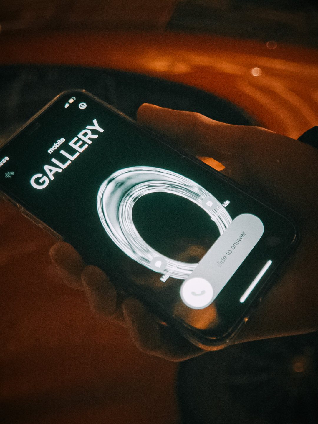 A phone in hand against the background of a dark orange car. The screen shows an incoming call labelled “GALLERY”. Dim, muted atmosphere.