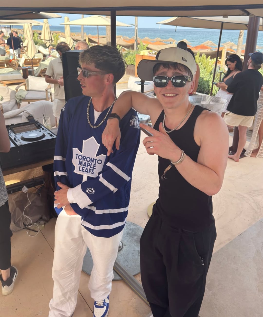 Tom Did It in a hockey jersey with GW beside the DJ decks at a daytime beach party by the sea.