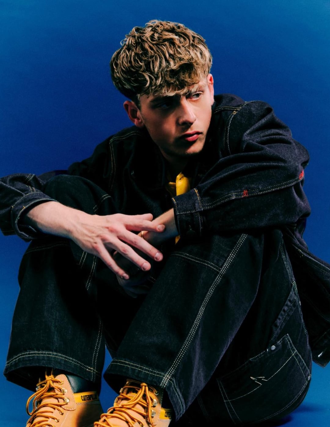 Tom Did It crouched in a denim jacket and worker boots against a deep blue backdrop.