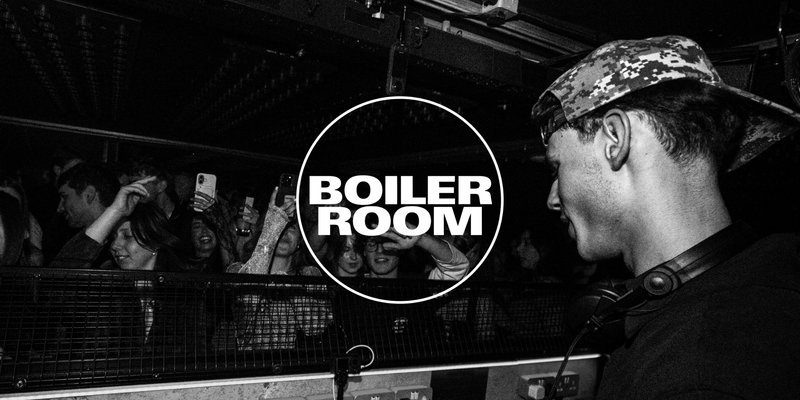 A DJ wearing headphones at a Boiler Room party. A crowd surrounds him, with many people filming on their phones. Energetic and lively mood. Black-and-white photo.