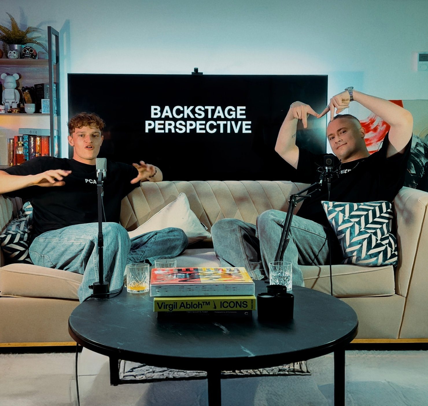 MNEEMO (Yaroslav Gorovoy) & MJP (Matthew John Potter) sitting on a sofa, speaking into microphones. The screen shows the text “Backstage Perspective”. A book and glasses are on the table. Cosy setting.