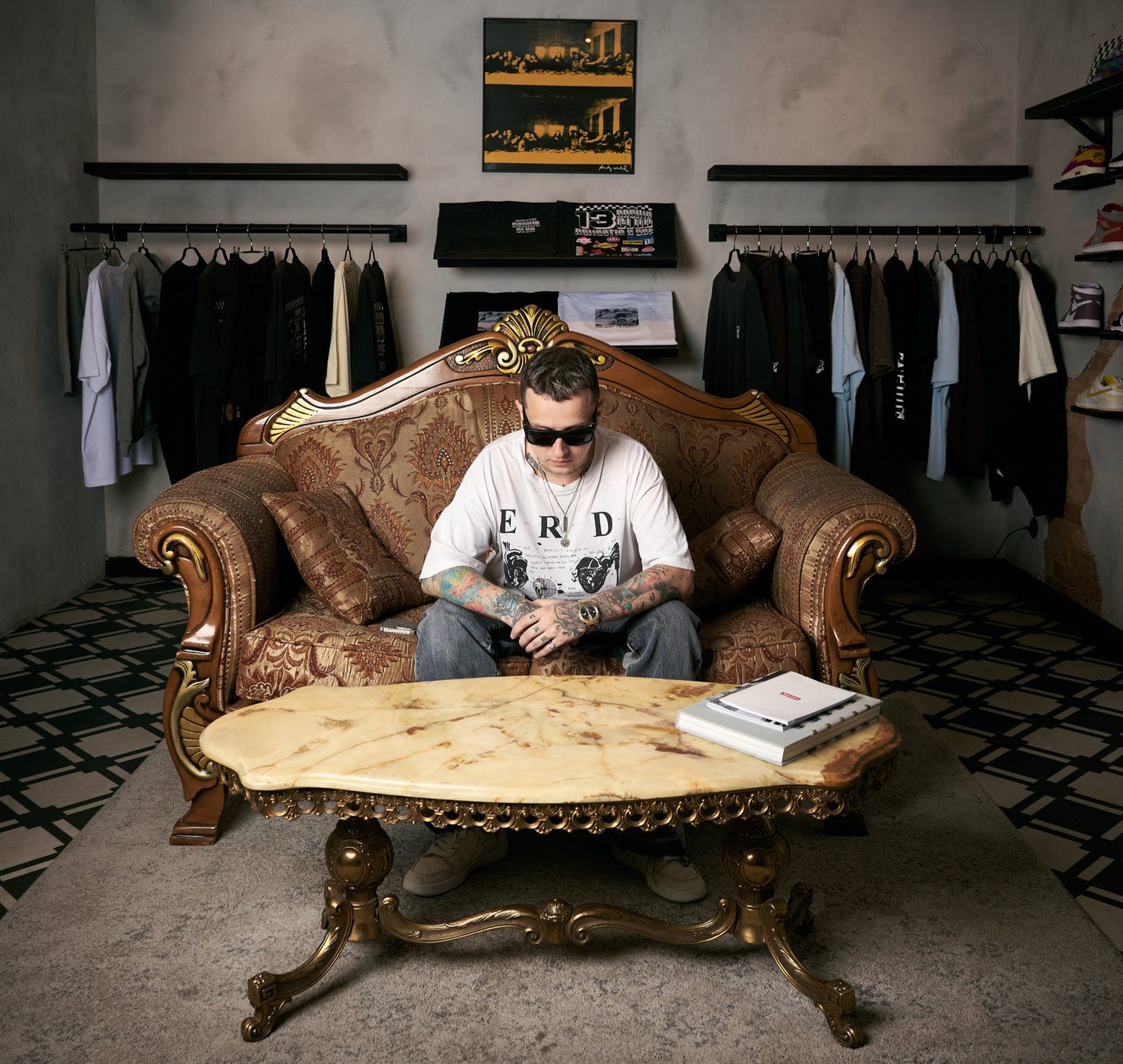 NIKITAIDISYUDA wearing sunglasses, sitting on a brown sofa with folded hands. Clothing racks behind him, and a marble table in front.