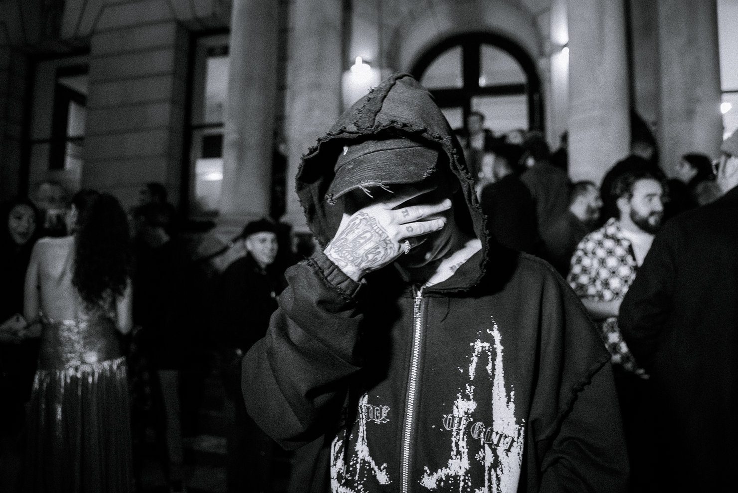 NIKITAIDISYUDA wearing a hood and cap, covering his face with a tattooed hand. Evening event, people in the background, black-and-white photo.