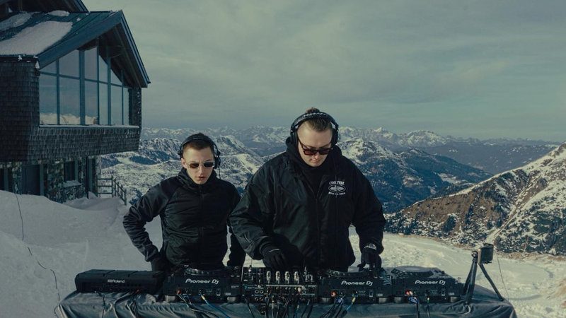 MNEEMO (Yaroslav Gorovoy) & SLAVKO (Viacheslav Zazymko) wearing headphones, performing in the snow in the mountains. In the background, there is a mountain landscape and a house with windows.